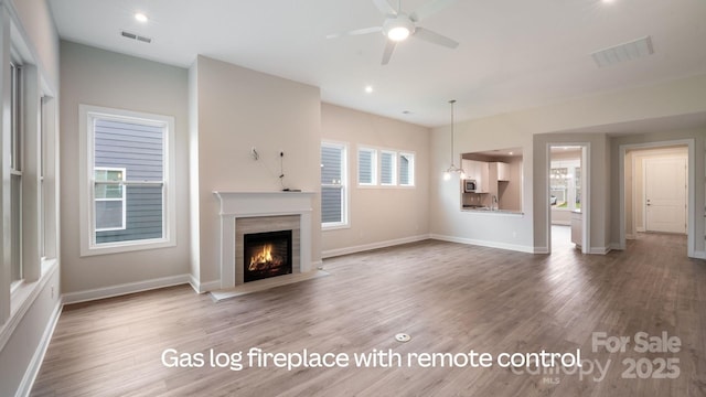 unfurnished living room with a warm lit fireplace, wood finished floors, visible vents, and a ceiling fan