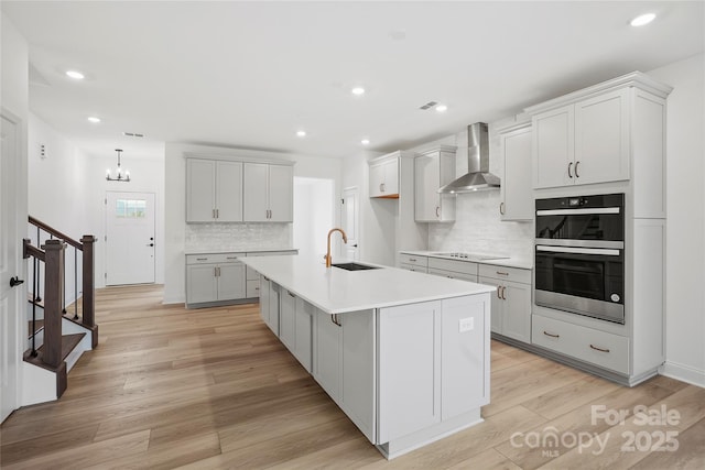 kitchen with black electric stovetop, light wood-style flooring, double oven, a sink, and wall chimney range hood