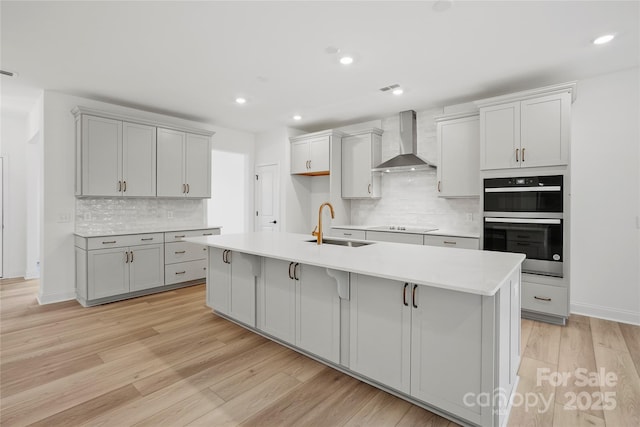 kitchen featuring gray cabinets, multiple ovens, light wood-style floors, a sink, and wall chimney range hood