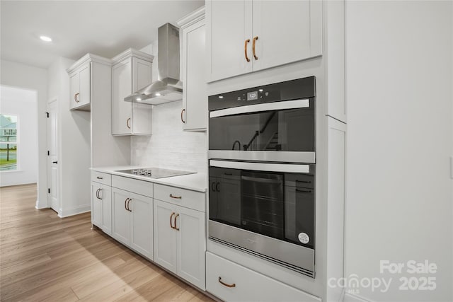 kitchen with double wall oven, black electric stovetop, light wood finished floors, light countertops, and wall chimney range hood