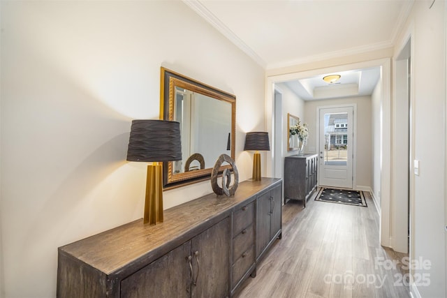 doorway with crown molding, a tray ceiling, baseboards, and light wood-style floors