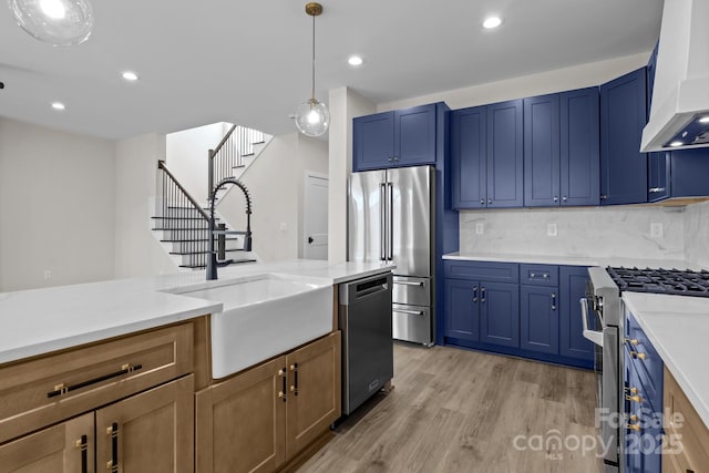 kitchen with a sink, high quality appliances, light wood-style floors, blue cabinetry, and wall chimney exhaust hood