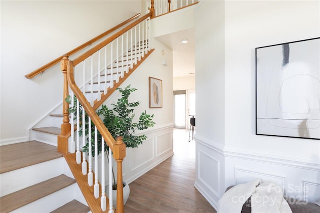 stairs with a high ceiling, a decorative wall, wood finished floors, and wainscoting