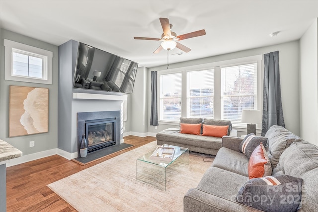 living area featuring a glass covered fireplace, ceiling fan, baseboards, and wood finished floors