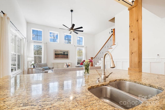 kitchen with light stone counters, a decorative wall, a sink, a ceiling fan, and wainscoting