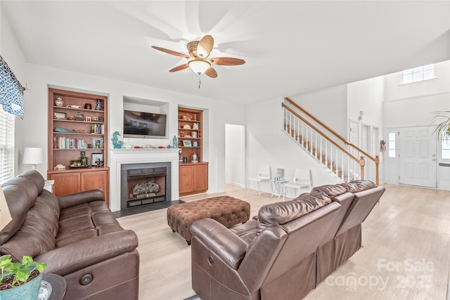 living room with ceiling fan, light wood-style flooring, a fireplace with flush hearth, built in features, and stairway