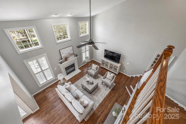 living area featuring a towering ceiling, a fireplace with flush hearth, a ceiling fan, wood finished floors, and baseboards