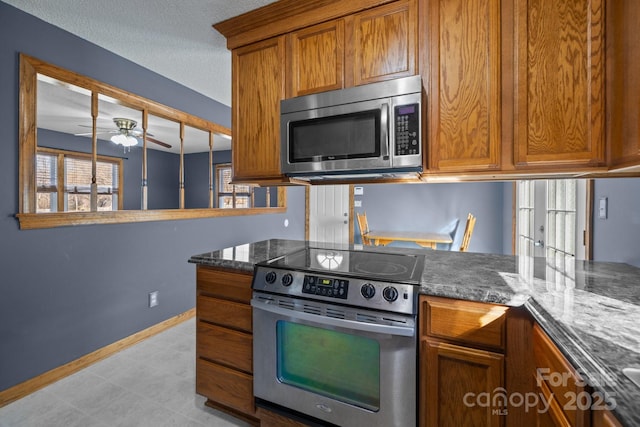 kitchen featuring appliances with stainless steel finishes, brown cabinetry, ceiling fan, dark stone countertops, and baseboards