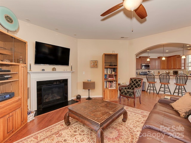 living room with arched walkways, a fireplace with flush hearth, visible vents, wood finished floors, and a ceiling fan