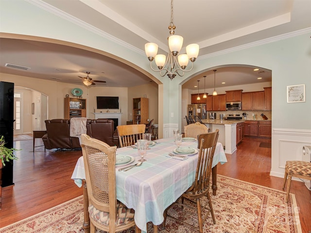 dining space with ceiling fan with notable chandelier, a fireplace, wood finished floors, visible vents, and a tray ceiling