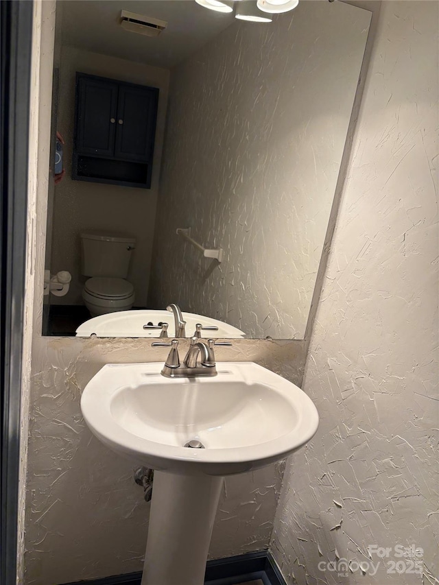 half bath with a sink, visible vents, toilet, and a textured wall