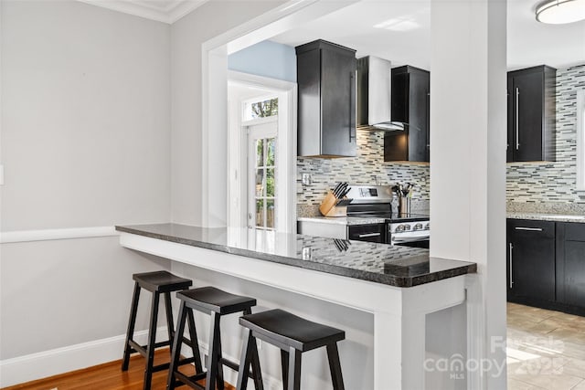 kitchen featuring dark cabinets, wall chimney range hood, stainless steel electric stove, tasteful backsplash, and a kitchen bar