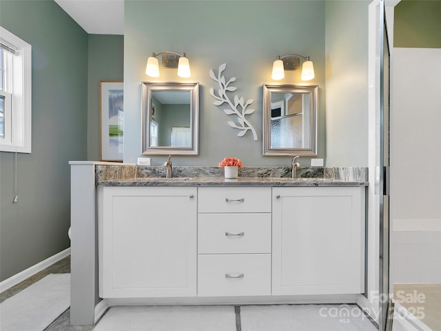 full bath with a tile shower, double vanity, a sink, and baseboards