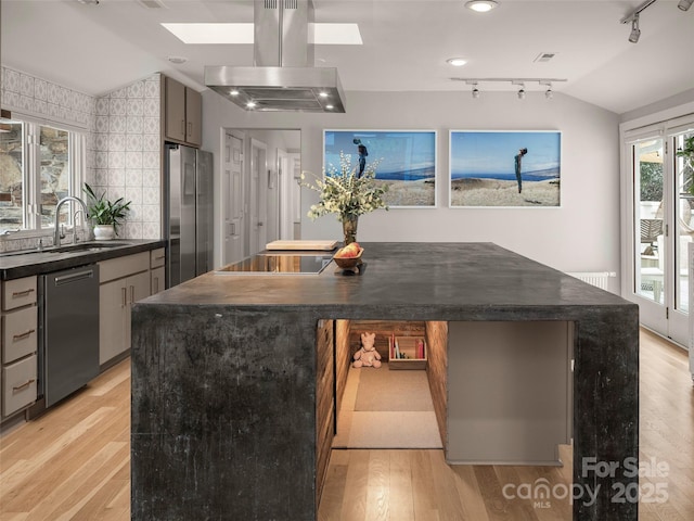 kitchen featuring island exhaust hood, stainless steel appliances, dark countertops, a sink, and vaulted ceiling with skylight