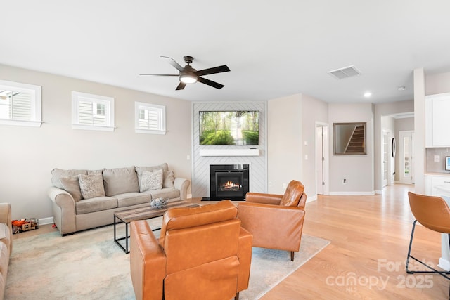 living area with light wood finished floors, visible vents, a ceiling fan, a large fireplace, and baseboards