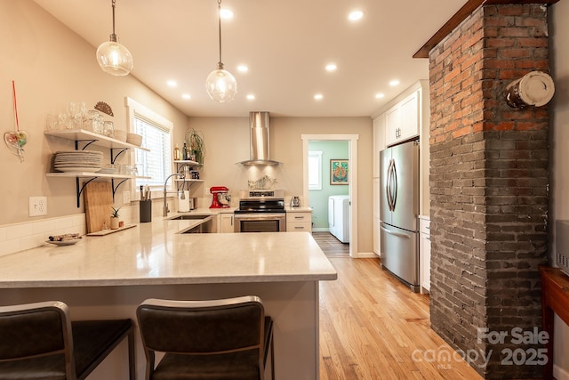 kitchen featuring appliances with stainless steel finishes, a peninsula, wall chimney range hood, open shelves, and a sink