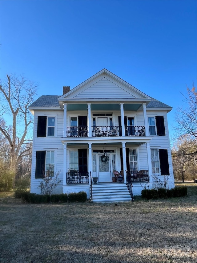 greek revival house featuring covered porch, a chimney, a front yard, and a balcony