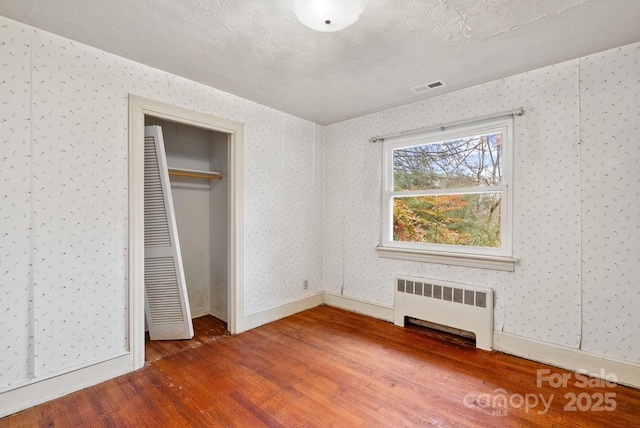 unfurnished bedroom with wood finished floors, visible vents, wallpapered walls, radiator heating unit, and a textured ceiling