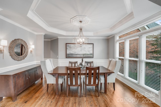 dining space with wainscoting, light wood-type flooring, an inviting chandelier, a raised ceiling, and crown molding