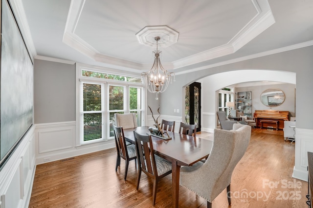 dining space featuring a tray ceiling, arched walkways, a wainscoted wall, light wood-style flooring, and a chandelier