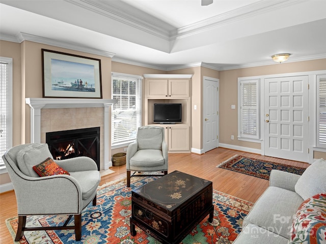 living area with baseboards, a tiled fireplace, light wood-style flooring, ornamental molding, and a tray ceiling