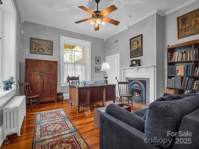 office area with ornamental molding, a ceiling fan, a warm lit fireplace, wood finished floors, and radiator heating unit