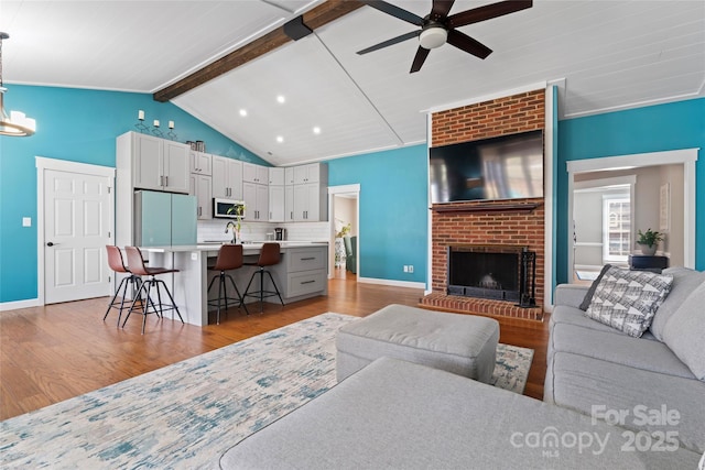 living area with vaulted ceiling with beams, light wood-style flooring, a brick fireplace, baseboards, and ceiling fan with notable chandelier