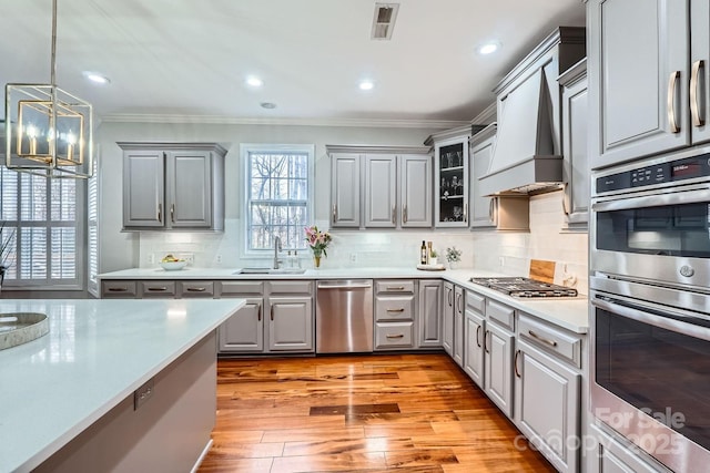 kitchen with gray cabinetry, custom range hood, a sink, appliances with stainless steel finishes, and crown molding