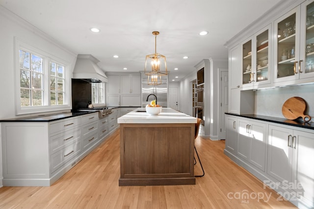 kitchen with tasteful backsplash, a center island with sink, custom range hood, glass insert cabinets, and light wood-style floors