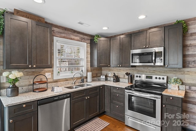 kitchen with light wood finished floors, appliances with stainless steel finishes, a sink, dark brown cabinetry, and light stone countertops