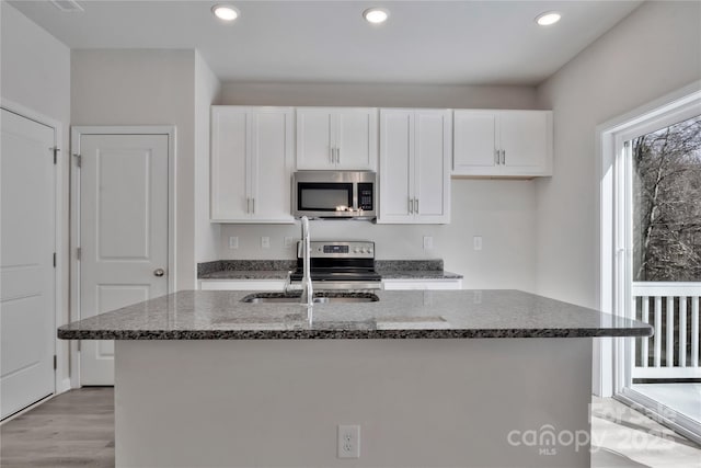 kitchen with stainless steel appliances, a sink, white cabinetry, an island with sink, and dark stone countertops