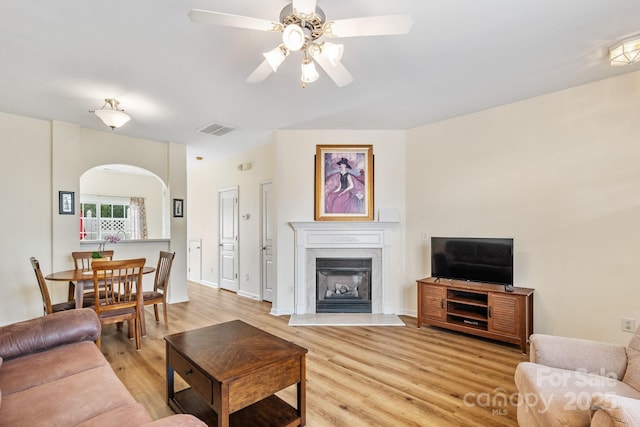 living area featuring ceiling fan, arched walkways, visible vents, light wood-style floors, and a glass covered fireplace