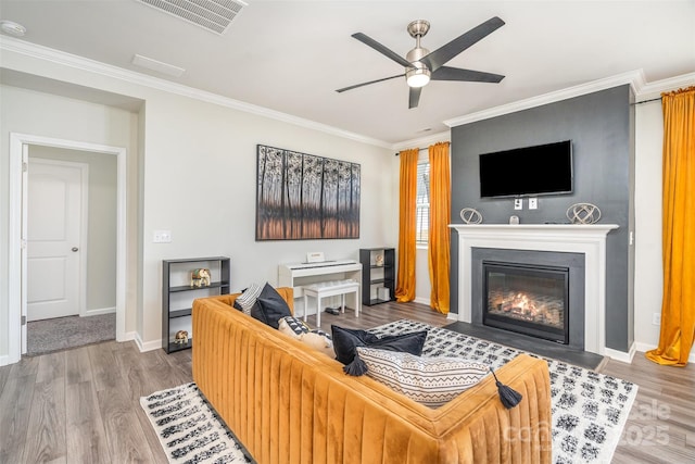 living room with a ceiling fan, wood finished floors, visible vents, a fireplace with flush hearth, and ornamental molding