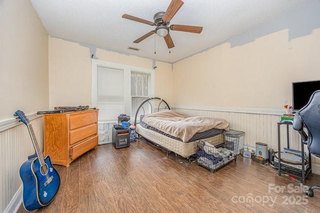 bedroom featuring a textured ceiling, a ceiling fan, wood finished floors, and wainscoting