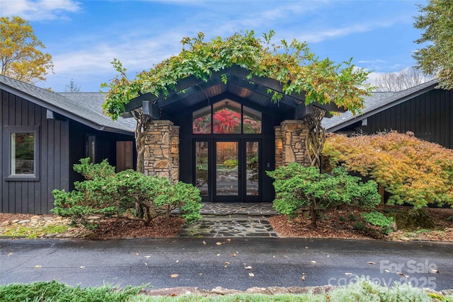 view of exterior entry featuring board and batten siding, stone siding, roof with shingles, and french doors