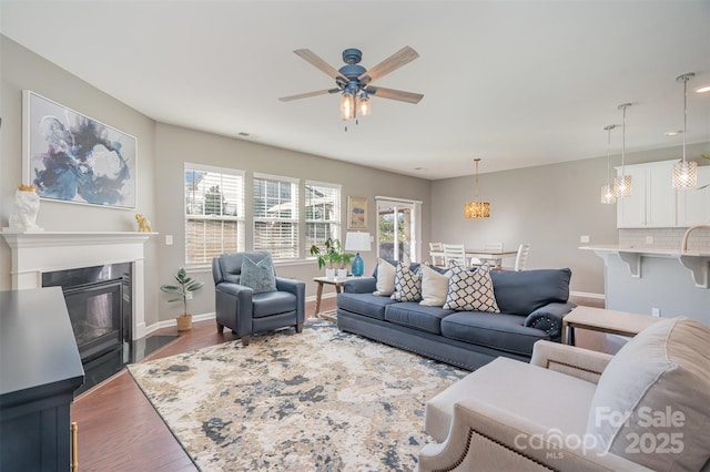 living area featuring visible vents, baseboards, a fireplace with flush hearth, wood finished floors, and ceiling fan with notable chandelier