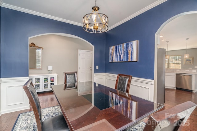 dining area with arched walkways, wood finished floors, wainscoting, an inviting chandelier, and crown molding