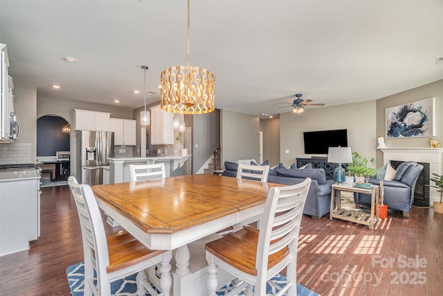 dining space featuring arched walkways, ceiling fan with notable chandelier, a fireplace, stairway, and dark wood finished floors