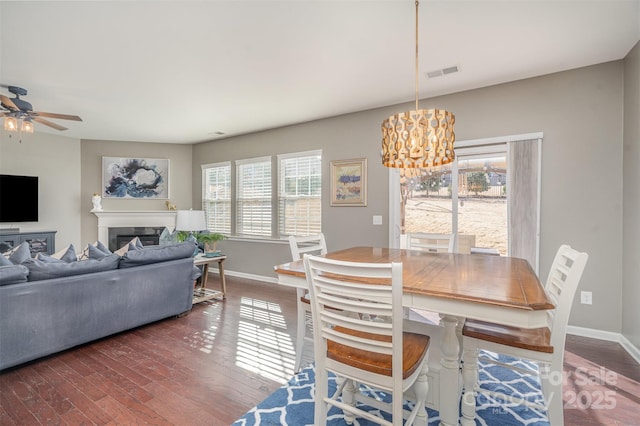 dining room featuring dark wood-style floors, a fireplace, visible vents, baseboards, and ceiling fan with notable chandelier