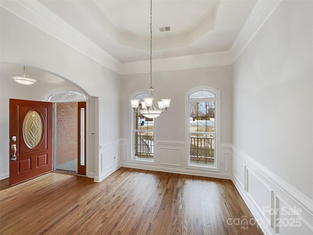 unfurnished dining area featuring an inviting chandelier, a raised ceiling, wood finished floors, and wainscoting