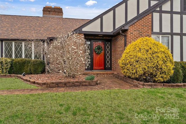 view of exterior entry featuring brick siding, roof with shingles, a chimney, stucco siding, and a lawn