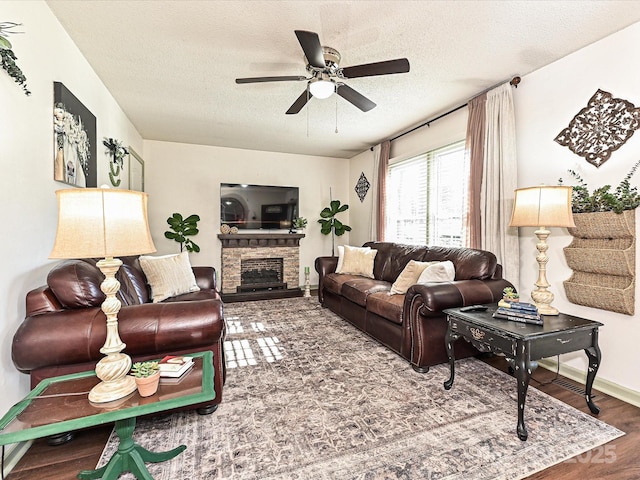 living area featuring wood finished floors, baseboards, ceiling fan, a stone fireplace, and a textured ceiling