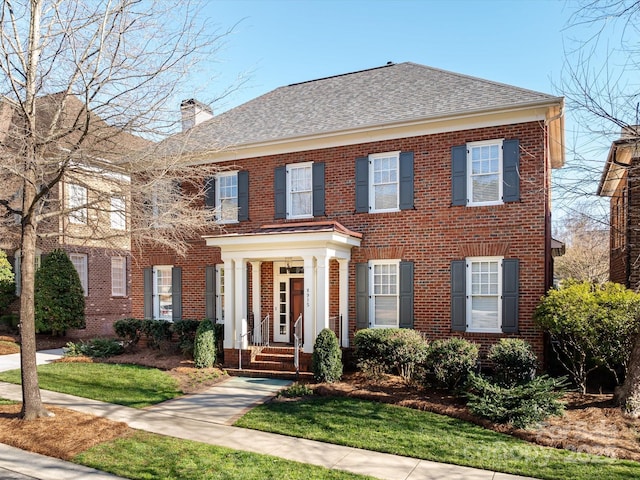 colonial inspired home featuring a shingled roof, brick siding, and a chimney