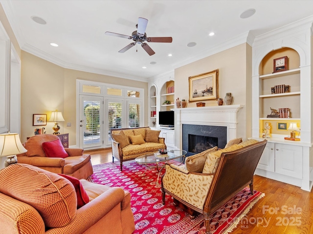 living area with ceiling fan, light wood-style flooring, a fireplace, built in features, and ornamental molding