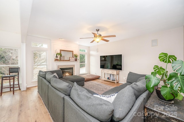 living area with a lit fireplace, baseboards, light wood-style flooring, and a ceiling fan