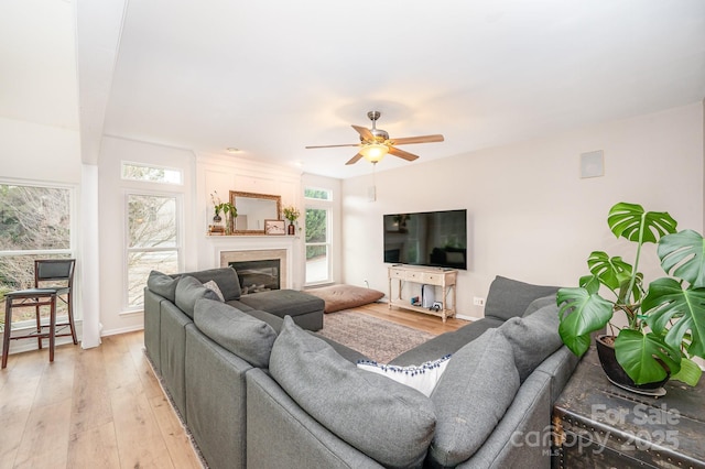 living area with ceiling fan, a glass covered fireplace, light wood-style flooring, and baseboards