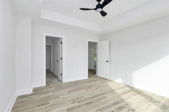 unfurnished bedroom featuring a walk in closet, baseboards, light wood-style flooring, a raised ceiling, and a ceiling fan