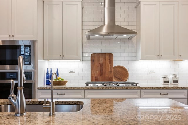 kitchen featuring decorative backsplash, appliances with stainless steel finishes, wall chimney exhaust hood, and light stone countertops