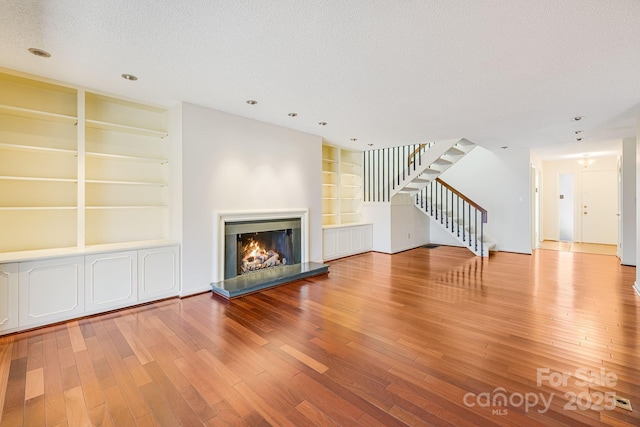 unfurnished living room with built in shelves, a textured ceiling, wood finished floors, a lit fireplace, and stairs