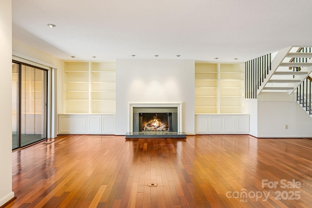 unfurnished living room with stairway, built in features, wood-type flooring, and a warm lit fireplace